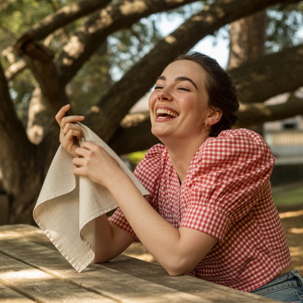 Model in red gingham blouse laughing at a sun-dappled picnic table, linen napkin in hand